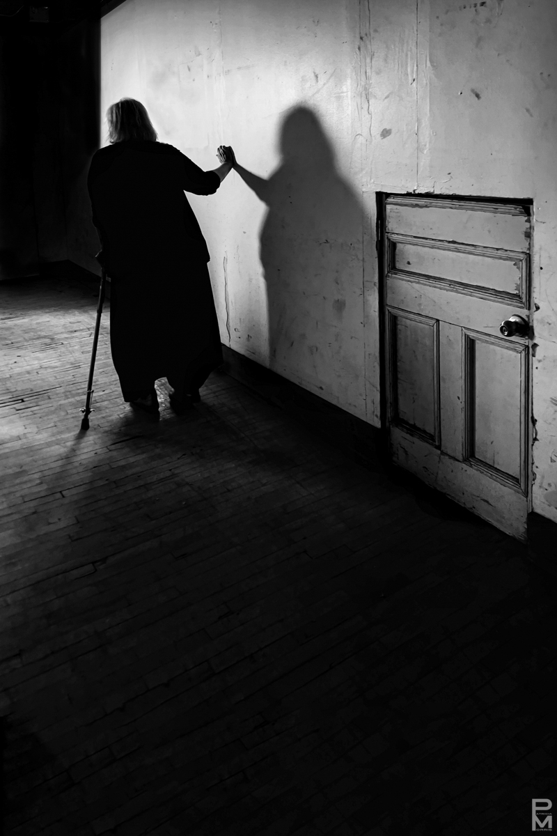 Digital photograph of the silhouette of an elderly, infirm woman with a cane walking down a hallway passing a mysteriously small door