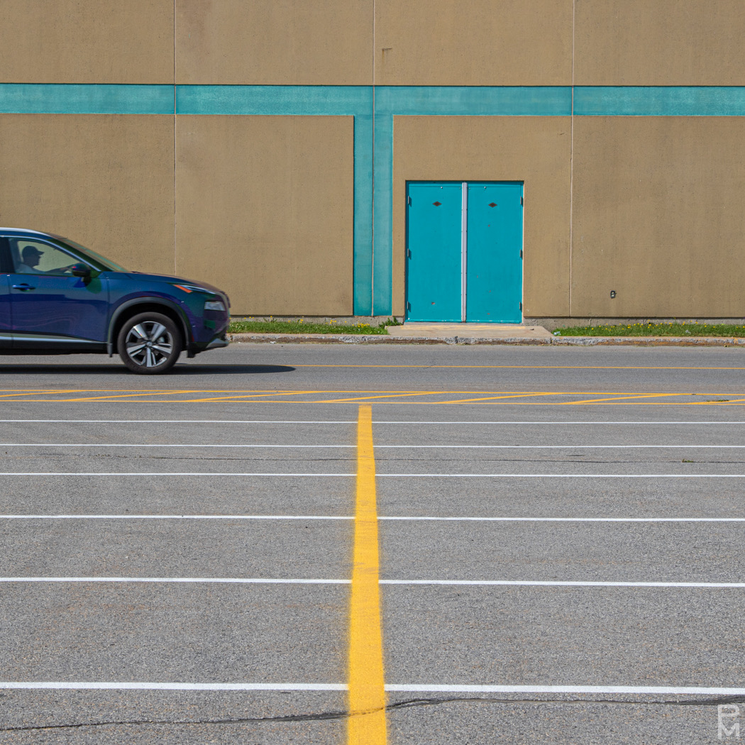 Digital photograph, minimalist image that includes a brick wall with door, blue car and parking lot markings