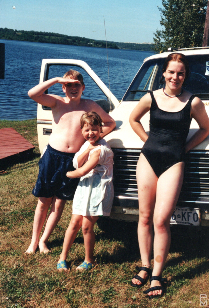 Snapshot of two young boys and a young woman in swimsuits by a white van at the water's edge