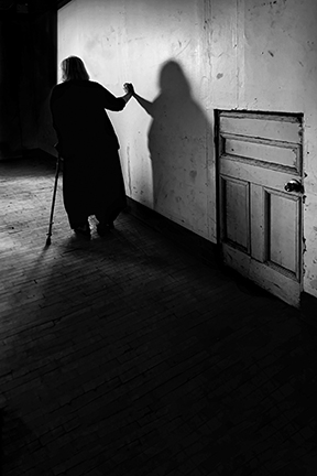 Black and white photograph of a woman with a cane walking down a hallway, her shadow cast on the wall beside an old door