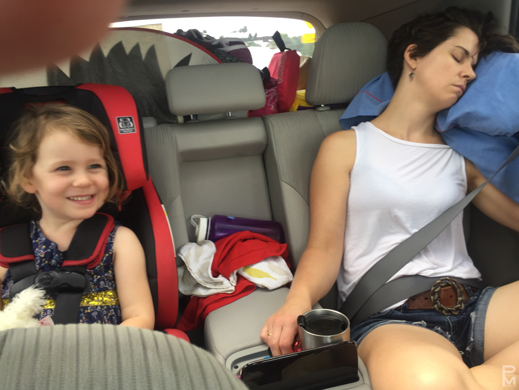 A child smiling in a car seat beside a sleeping passenger during a road trip
