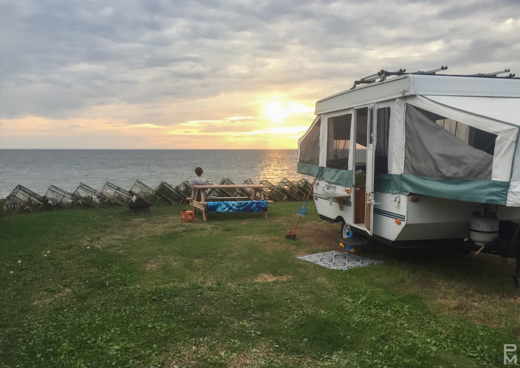 A pop-up camper trailer parked on a grassy bluff overlooking the ocean at sunset