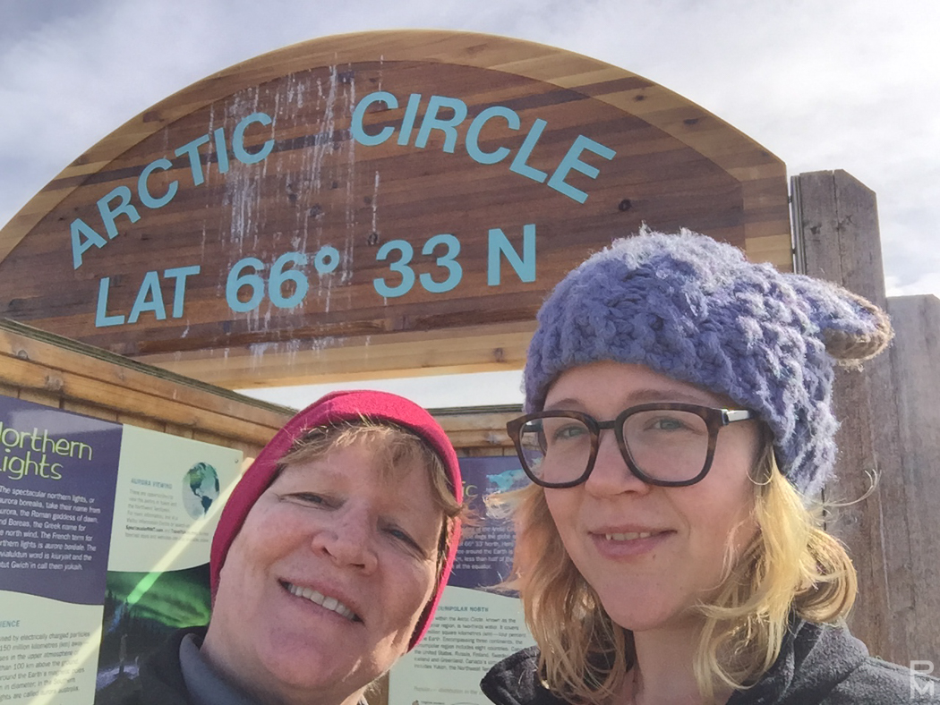 Two people posing under the Arctic Circle sign at latitude 66 degrees 33 minutes north