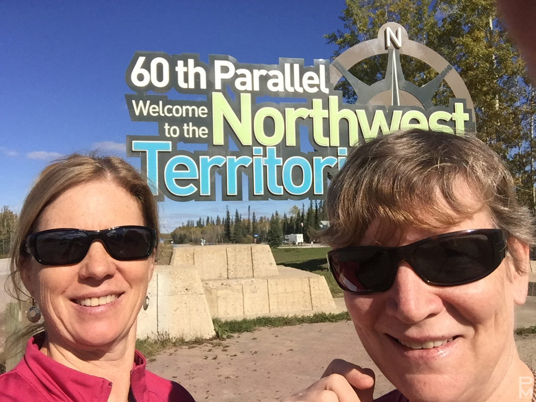 Two people posing at the 60th Parallel sign welcoming visitors to the Northwest Territories