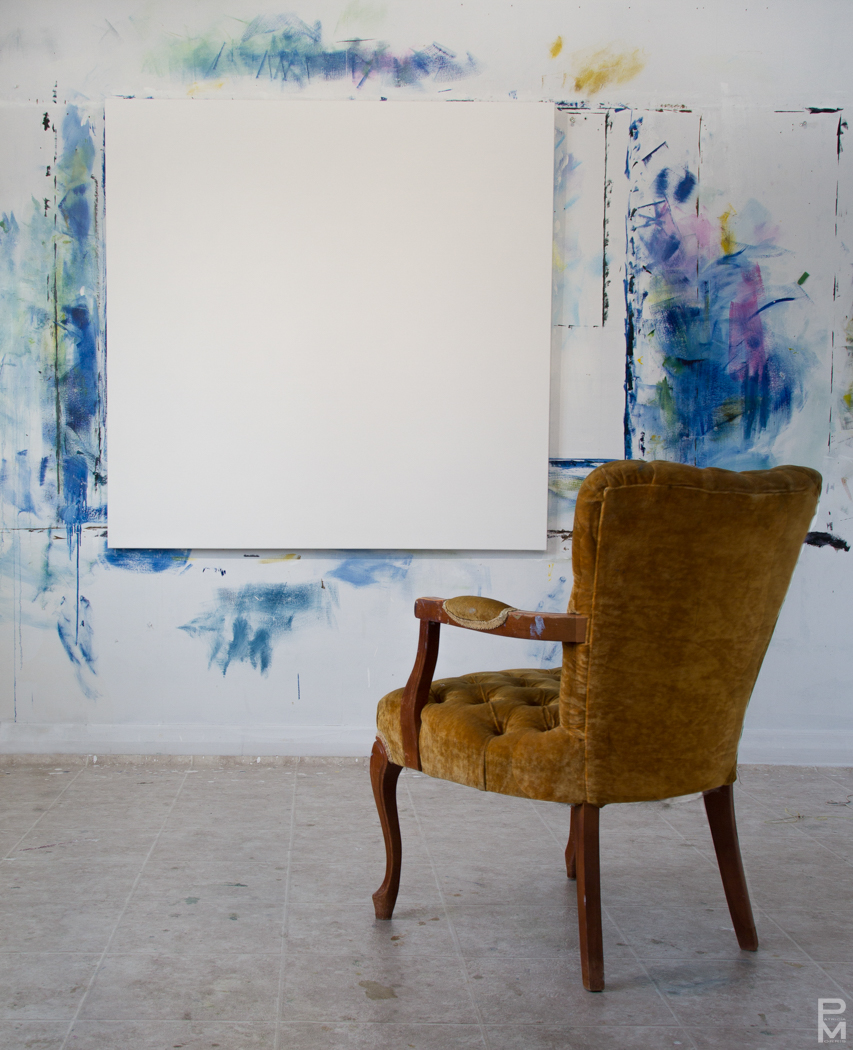 A blank canvas leaning against a paint-splattered studio wall next to an antique chair
